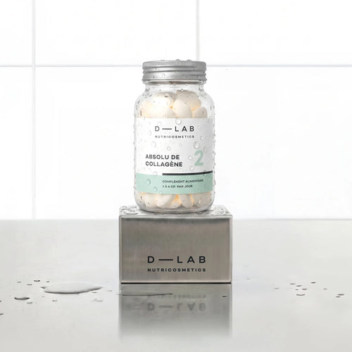 Absolu de Collagène D-LAB Nutricosmetics cure 1 mois, flacon en verre de comprimés perlé de gouttes d'eau posé sur un socle inox brossé, fond carrelage blanc
