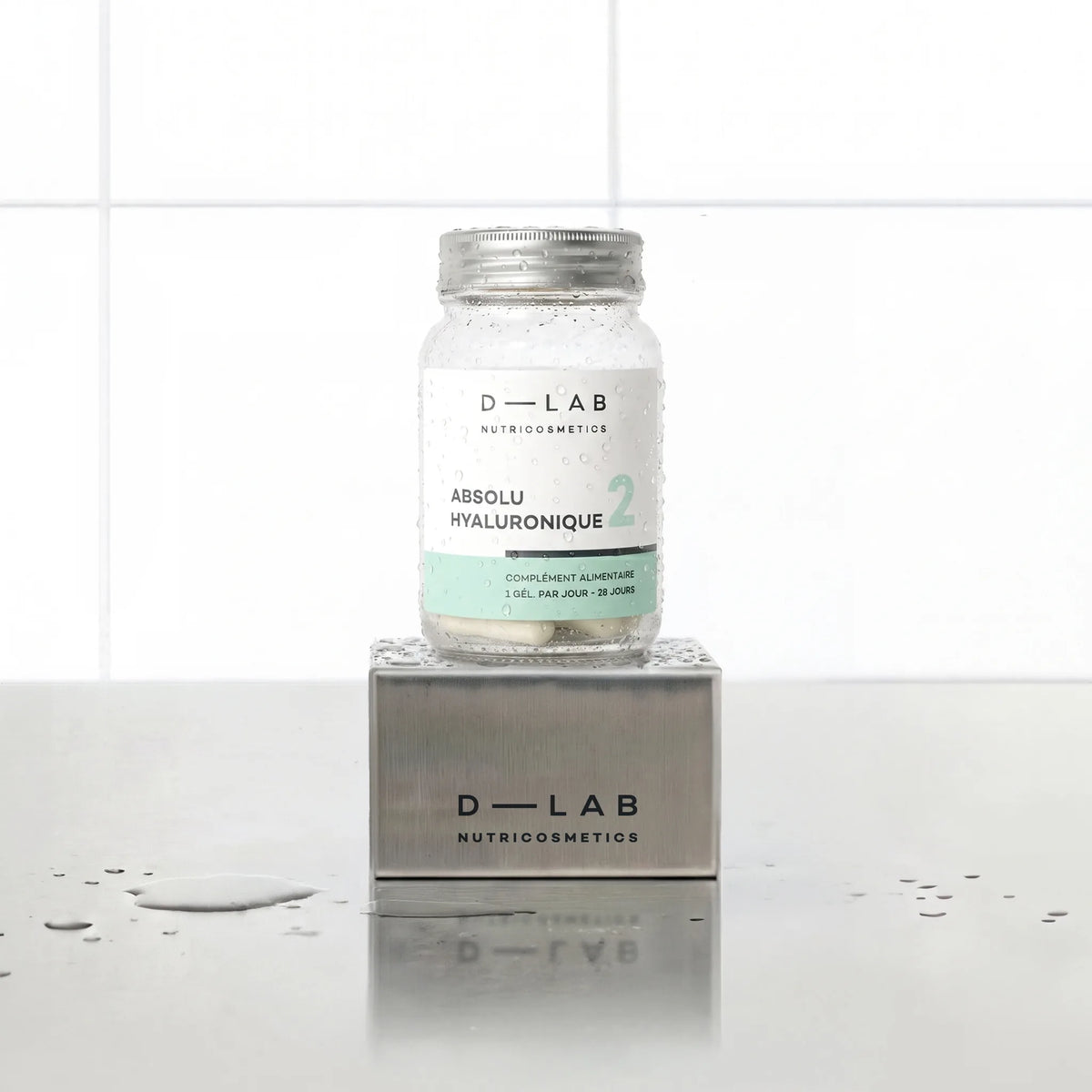 Absolu Hyaluronique D-LAB Nutricosmetics, cure 1 mois : flacon en verre de gélules posé sur un socle inox brossé, fond carrelage blanc