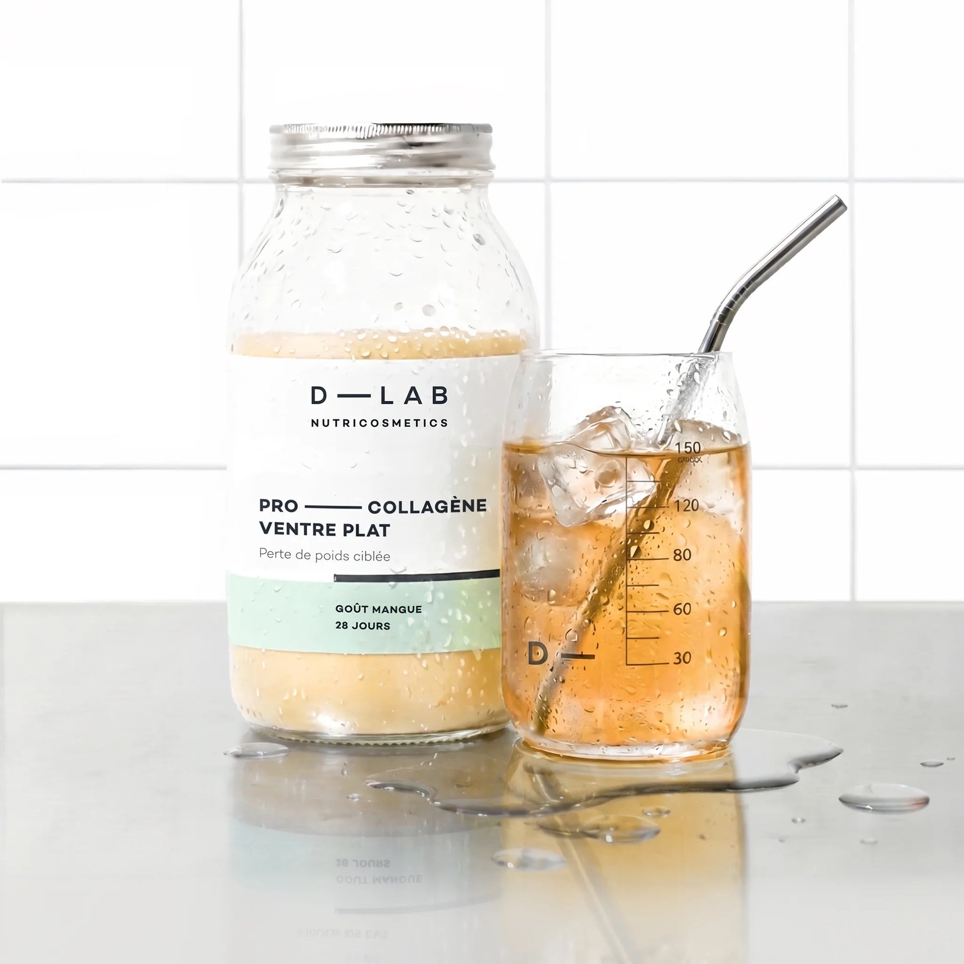 Flacon en verre Pro-Collagène Ventre Plat goût mangue D-LAB et verre glacé de boisson ambrée avec glaçons et paille inox, plan de travail carrelé blanc