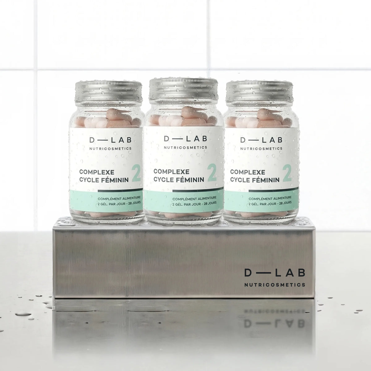 Complexe Cycle Féminin D-LAB Nutricosmetics, cure 3 mois : 3 flacons de gélules alignés sur un socle inox brossé, fond carrelage blanc