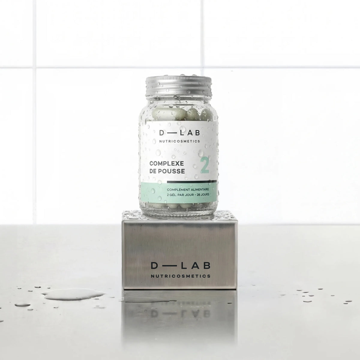 Complexe de Pousse D-LAB Nutricosmetics cure 1 mois, flacon en verre de gélules posé sur un socle inox brossé, fond carrelage blanc et gouttes d'eau
