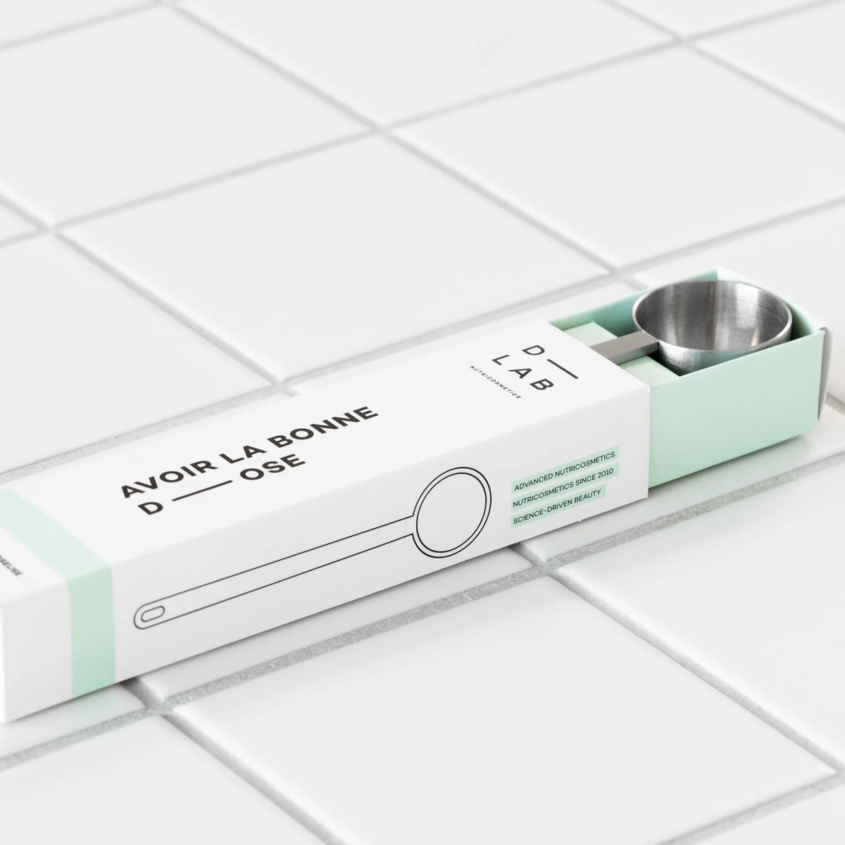 Cuillère doseuse D-LAB en inox glissée dans son étui blanc et vert d'eau « Avoir la bonne dose », posée sur un carrelage blanc