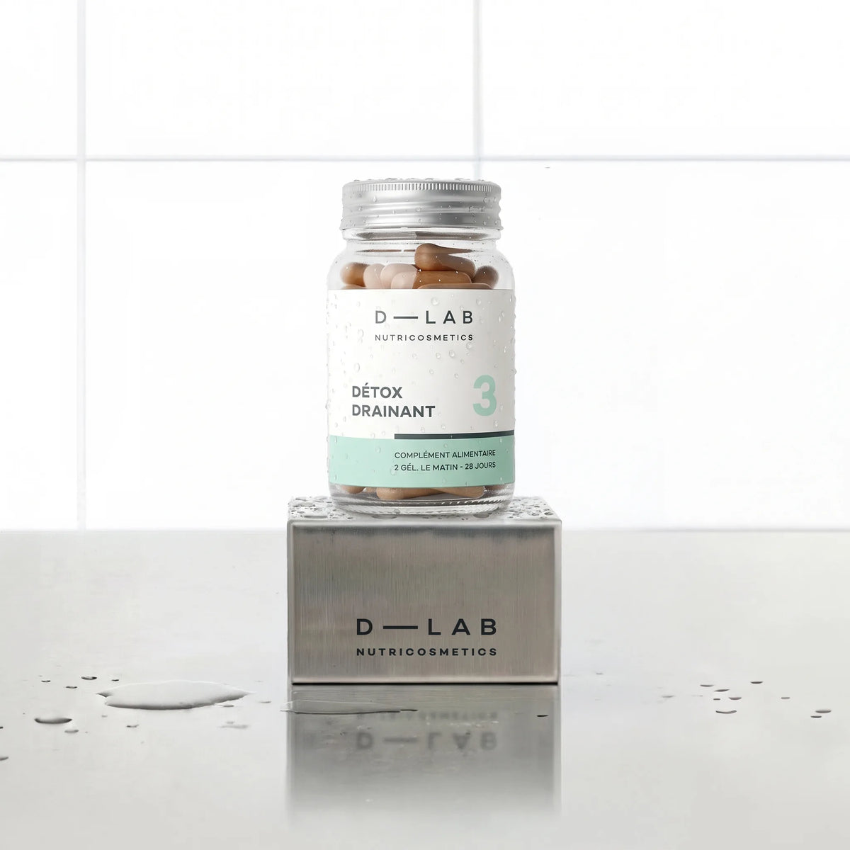 Détox Drainant D-LAB Nutricosmetics, cure 1 mois : flacon en verre de gélules posé sur un socle inox brossé, fond carrelage blanc