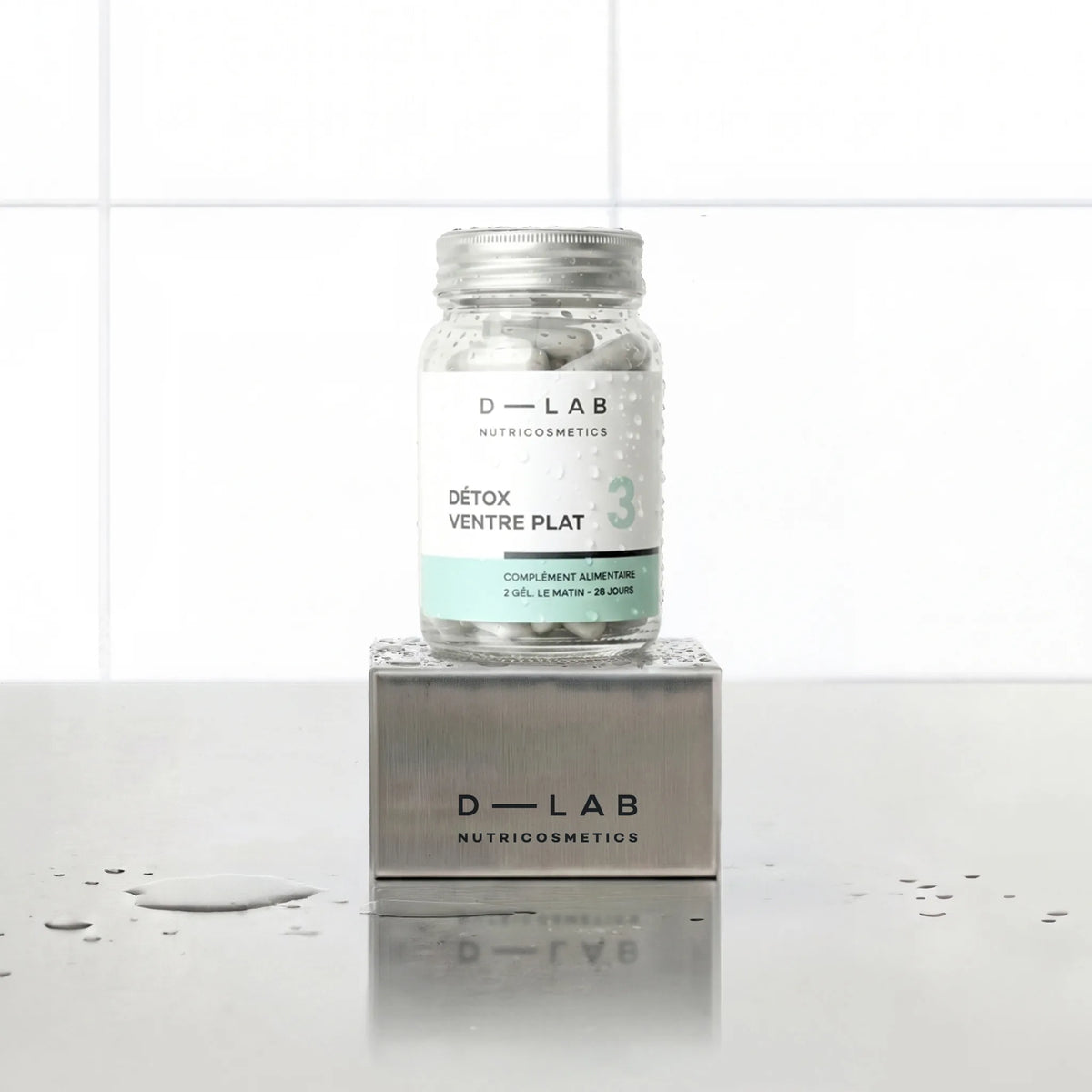 Détox Ventre Plat D-LAB Nutricosmetics, cure 1 mois : flacon en verre de gélules posé sur un socle inox brossé, fond carrelage blanc